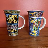 Two Dunoon fine stoneware mugs with colorful Celtic knotwork and animal motifs on wood surface against pink wall.