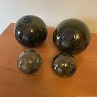 Four dark green glass fishing net floats shown on table, two larger and two smaller sizes, each with a seal mark on top.