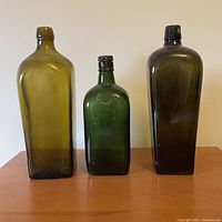Three green antique glass bottles of varying heights and shapes placed side by side on a wooden surface against a plain wall.