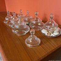 Seven short, wide apothecary jars with lids displayed on a wooden surface, some filled with shells and decorative items, one extra lid in front.