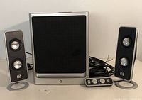 Front view of two black satellite speakers with silver trim, front of black subwoofer with silver frame, and wired remote control