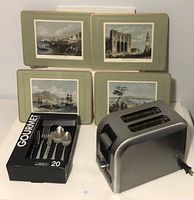 Boxed 20-piece stainless steel flatware set by Safdie, next to Oster stainless steel toaster, with four vintage style placemats in the background