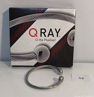 Front view of Q Ray twisted silver-tone wellness bracelet with two round end caps in front of original branded box.