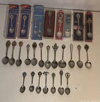 Top view of 27 vintage collectible spoons, some in original branded packaging and many loose spoons with detailed heraldic handle designs.