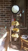 Full view of black metal tall stand with round clock face and three plant holders filled with faux sunflowers against brick wall background