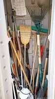 Photo showing assortment of garden tools stored inside small shed, including multiple shovels, rakes, brooms, and a sprayer