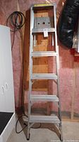 Full view of the closed aluminum step ladder leaning against a wall in a workshop setting.