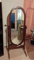 Full view of wood framed oval cheval mirror standing in basement room, showing dark wood frame and side supports.