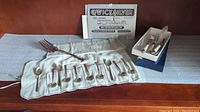 Ten International sterling coffee spoons laid out on a cloth roll, silver plate serving fork placed above them, cheese utensils in a box and Quick Silver cleaning plate behind.