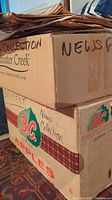 Two cardboard boxes labeled 'News Collection' containing vintage newspapers, one labeled B.C. Apple boxes used for storage.