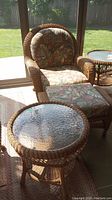 Front view of wicker armchair with floral cushions, matching ottoman and round side table under glass