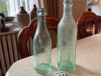 Two antique aqua glass bottles with embossed brewery names placed on a white tablecloth indoor setting.