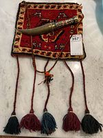 Photo showing carpet bag with traditional Persian design and five tassels, along with ceremonial cutting utensil with ornate handle and curved sheath