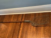 Full view of the antique potato digger tool showing the long wooden handle and metal prongs head, placed on a wooden floor.