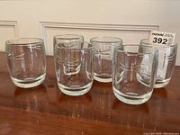 Six clear glass tumblers displayed in a row on a wooden surface. Each has a thick base and clear sides with a dragonfly design. This shows the overall quantity and condition.