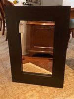 Front view of a rectangular mirror with a black wood grain frame placed on a carpeted floor reflecting furniture.