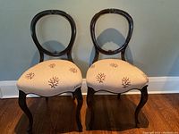 Full view of the two balloon back chairs side by side, showing the dark wood frames and coral-patterned beige upholstered seats. Both chairs have Queen Anne style curved legs.
