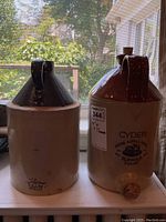 Two 1 gallon stoneware crocks side by side, one tan and brown glazed, the other with an off-white bottom and dark brown top and handle.