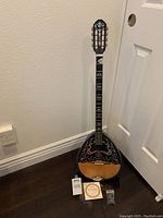 Full view of 8 string Greek bouzouki standing against wall with stand, showing body, neck, and headstock with inlay design. Picks and extra strings placed near base.