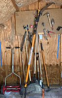 Photo of various garden tools leaning against a wooden wall and pegboard, showing a pole trimmer, rake, hoe, push mower, and other tools.