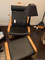 Full view of IKEA POANG armchair with matching footrest and black table lamp positioned beside the chair on dark hardwood floor.