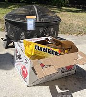 Metal fire pit with a box containing Duraflame firelog and wood poker in front of it on concrete ground