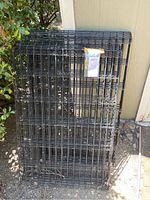 Folded stack of black metal wire mesh panels for pet exercise pen, photographed outdoors near shrubbery and building wall.