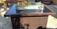 Brown faux wicker fire pit table with clear tempered glass top and central burner ring filled with glass beads