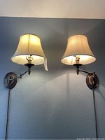 Pair of brass swing arm plug-in wall lamps mounted on wall, both lamps lit showing warm light through fabric shades