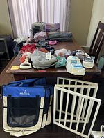 Multiple small dog supplies laid out on a table including dog clothes, potty pads, treats, carrier, and pet gate on the floor in front of the table