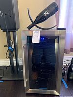 Wine fridge with glass door, digital display showing temperature, and stainless steel handle. Balancing wine holder on top holding one wine bottle at an angle.