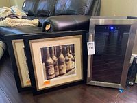 Overall shot showing two framed lithographs leaning against a black leather couch and a stainless steel wine fridge with glass door and digital display