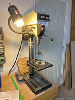 Full side view of the Delta drill press on a workbench showing the lamp light attached, base, and worktable with accessories on top.