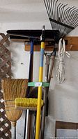 Photo showing various long handled yard tools hanging on a wall, including brooms, rakes, spade, hoe, and an extension pole.