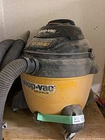 Photo of the front left side of the wet/dry vac showing yellow tank, black top housing, hose attached to the front and labeled 'shop-vac ultra'. Dusty condition visible.