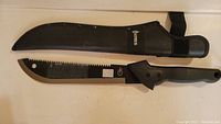 Gerber machete next to its protective black sheath on a white surface.