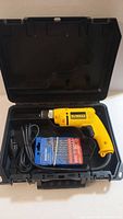 Open black plastic carrying case showing the yellow DeWalt electric drill, blue pack of drill bits, and power cord coiled inside.