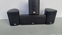 Four JBL speakers arranged with three satellite speakers and one center channel speaker, all black with metal grilles and JBL logos visible.