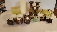 Photo showing green and off-white pillar candles, spherical white candles, wooden candle holders, brass vases, and figurines.