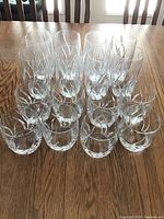 Photo shows 16 clear Mikasa glasses arranged on a wooden table. Front row features 8 short tumblers, back row features 8 tall glasses. Both glass types are clear with etched diagonal lines as decoration.