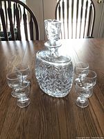 Front view of cut glass decanter surrounded by six small glasses on a wooden table.