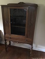 Front view of old wooden china cabinet, showing closed glass center door flanked by two side wooden panel doors, drawer with two knobs, and turned legs.