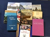 Books and flags arranged on a dark cloth background, showing titles related to Kingston and Prince Edward County history plus five vintage British flags on wooden poles.