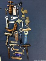 Top-down view of collection of 22 old bottle openers and corkscrews arranged on a blue cloth background. Various designs, materials, and branding visible.