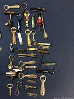 Top-down view of entire lot showing variety of vintage bottle openers and corkscrews in different materials, shapes, and sizes spread on dark surface.