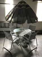 Photo showing full patio set with glass table, umbrella, and four chairs in indoor setting.
