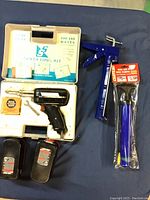 Top-down view of the soldering kit case open showing soldering gun and two Black & Decker batteries, blue caulking dispenser, and packaged purple bicycle mini-pump on blue fabric surface.