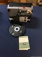 Sankyo Super 8 dual format projector with controls, black plastic film reel, and instruction manual on a dark blue surface.