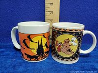 Front view of both mugs showing Halloween themed designs and measuring scale for height.