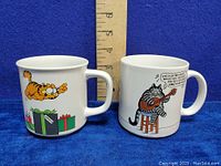 Photo showing both mugs side by side with a ruler for size reference. Garfield mug with cat jumping over gifts and B. Kliban cat playing guitar visible.